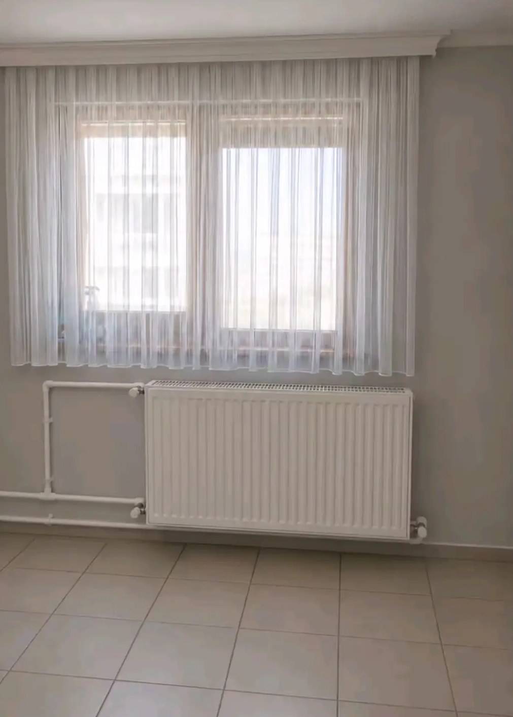 Pencere altında panel radyatör ve beyaz boyalı kalorifer boruları